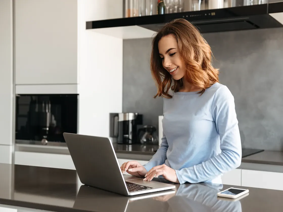 A woman smiling at her laptop at a kitchen counter, her phone beside it