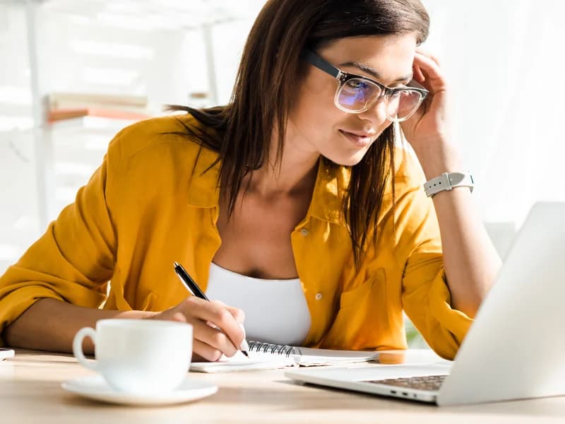 A woman writing notes at her desk beside a laptop